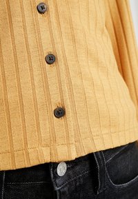 Senapsskalad stickad topp med mörkbruna knappar och en figurnära design, kombinerad med mörka denimjeans och synlig knappdetalj.