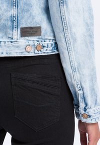 Jasnoniebieska kurtka dżinsowa z miedzianymi akcentami, zestawiona z czarnymi dżinsami, które mają tylną kieszeń z poziomymi szwami.