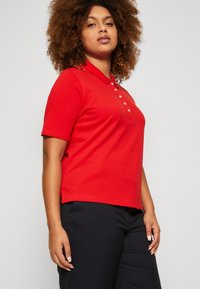 Camisa polo roja de algodón suave, con cuello y tapeta de tres botones, combinada con pantalones negros para un look casual.