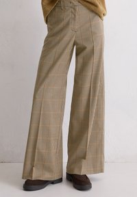 Brede bruine geruite pantalons gecombineerd met donkerbruine schoenen tegen een effen lichte achtergrond.