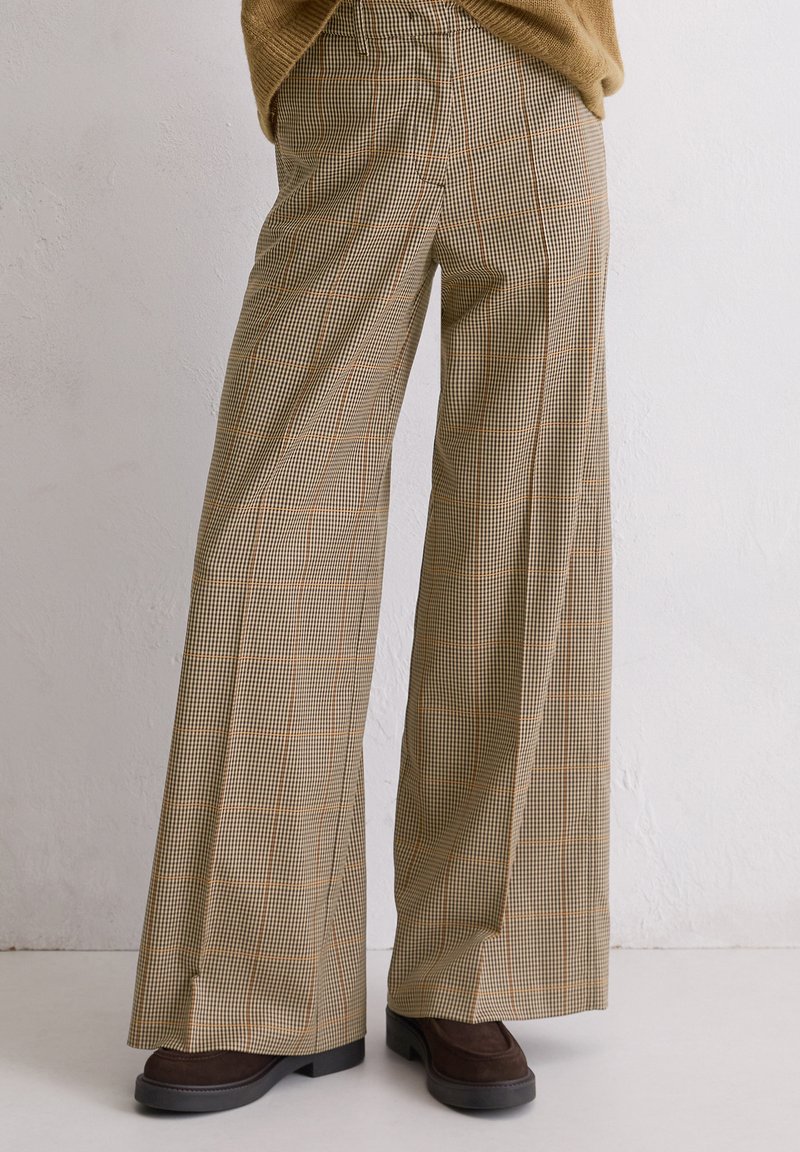 Brede bruine geruite pantalons gecombineerd met donkerbruine schoenen tegen een effen lichte achtergrond.