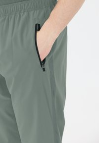 Lichte stretchbroek in een gedempte groene stof. Voorzien van een ritszak met zwarte afwerking. Elastische tailleband voor een verstelbare pasvorm.