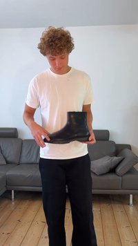 Botim de tornozelo preto em borracha com uma textura suave, bico arredondado e sola baixa, segurado por uma pessoa vestindo uma camisa branca e calças pretas.