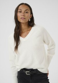 Elfenben v-hals sweater i blødt strik, med lange ærmer og ribbet kant. Styles med sort bælte over mørke jeans. Simpelt design, minimal tekstur.