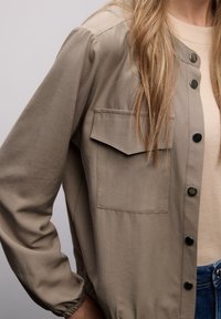 Veste légère marron clair à boutons, avec une poche poitrine, des poignets élastiqués et une texture lisse et mate, portée avec un haut beige et un jean bleu.