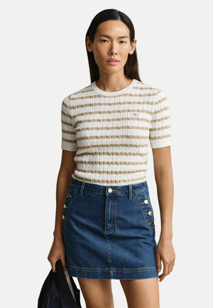 Vrouw draagt een beige-wit gestreepte korte mouw trui en een blauwe denim minirok met gouden knoopdetails, en houdt een donker jasje vast.