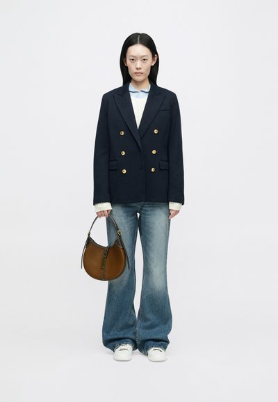 Blazer azul marinho de dupla abotoadura com botões dourados, suéter creme e camisa azul clara, combinado com jeans flare e uma mala castanha. Ténis brancos.