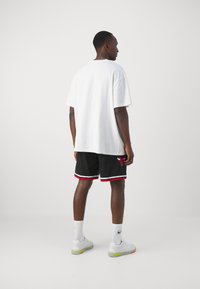 Maglietta bianca oversize, pantaloni sportivi neri con orlo a righe rosse e bianche e logo dei Chicago Bulls, abbinati a sneakers bianche e calze corte.
