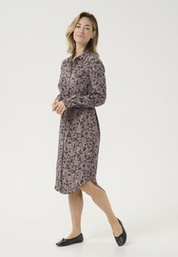 Lange mouwen, knielange jurk van zachte stof met een mauve basis en een zwart bloemenpatroon en knopen aan de voorkant, gecombineerd met zwarte flats.