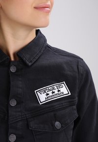 Giacca in denim nera con chiusura a bottoni, colletto a punta e una patch bianca con la scritta "VINTAGE RED ARTISTIC TOUCH" e tre stelle.