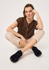 Haut tricot brun à boutons avec des manches courtes, pantalons cropped beiges et baskets en suede noires avec des rayures grises, posé assis avec les jambes croisées.