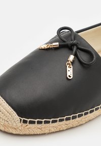 Melnas ādas espadrilles ar austu zāģēšanas zoli, kurai ir dekoratīva loka un zelta tonēts piekariņš sānos. Gluda tekstūra.