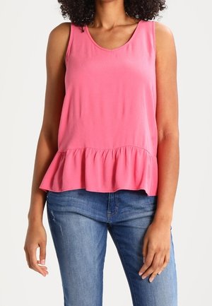 Vrouw die een mouwloze roze peplumtop en een blauwe spijkerbroek draagt, staand tegen een witte achtergrond met ontspannen armen.