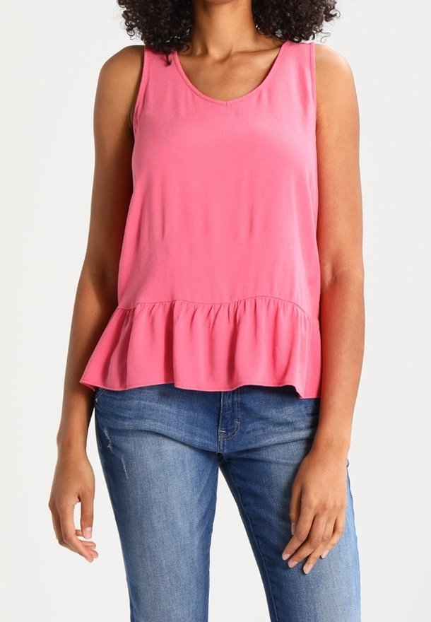 Femme portant un haut rose à basque sans manches et un jean bleu, debout contre un fond blanc avec les bras détendus.