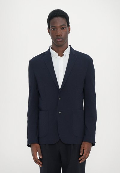 Blazer navy hecho de tela suave, de un solo botón con dos bolsillos frontales, llevado sobre una camisa blanca con cuello. Ajuste entallado, diseño minimalista.