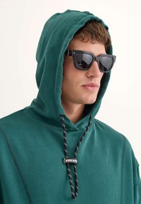 Sudadera verde con capucha y cordones, featuring cords de patrón en blanco y negro. Llevada con gafas de sol negras, destacando el rostro.