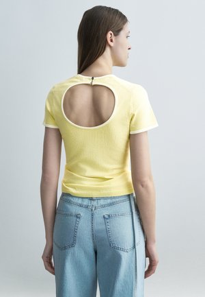 Mujer con cabello largo y castaño que lleva una camiseta amarilla de manga corta con un corte circular en la espalda y jeans de tiro alto azul claro.