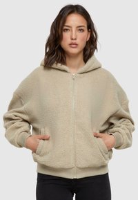Felpa beige in pile con zip frontale, vestibilità oversize, cappuccio regolabile e tasche frontali. Tessuto testurizzato con polsini e orlo a coste.