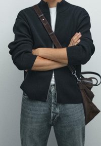 Henkilö, jolla on kädet ristissä, pukeutuneena mustaan vetoketjulliseen huppariin, harmaisiin farkkuihin ja ruskeaan olkalaukkuun, yksinkertaista taustaa vasten.