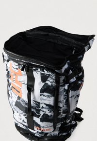 Αντικατάσταση πλάτης με ασπρόμαυρο σχέδιο και πορτοκαλί λεπτομέρειες. Διαθέτει πολλαπλές θήκες, δίχτυα και ανθεκτικά φερμουάρ. Το υλικό φαίνεται ανθεκτικό.