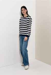 Raidallinen neulepaita, jossa on laivastonsinistä ja kermaa, yhdistettynä vaaleansinisiin levenevin farkkuihin. Malli seisoo yksinkertaisella taustalla ja jalassa on valkoiset tennarit.