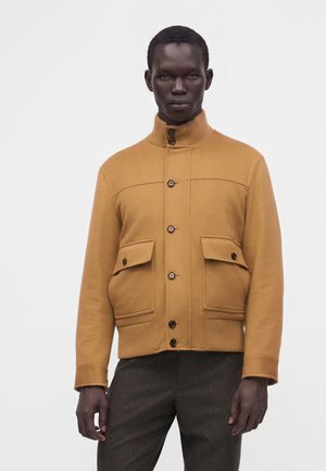 Mann trägt eine senfgelbe, geknöpfte Jacke mit hohem Kragen und zwei vorderen Klappentaschen, kombiniert mit dunkelgrauen Hosen.