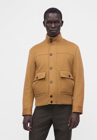 Homme portant une veste jaune moutarde boutonnée avec un col montant et deux poches à rabat à l’avant, associée à un pantalon gris foncé.