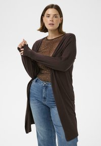 Langer, offener Cardigan in Dunkelbraun, mit langen Ärmeln und weichem Stoff, über einem transparenten Oberteil mit Punkten getragen, kombiniert mit blauen Jeans.