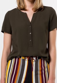 Blouse olive verte à manches courtes avec un décolleté en v et cinq boutons. Présente une texture lisse et une coupe décontractée, assortie à un pantalon à rayures multicolores.