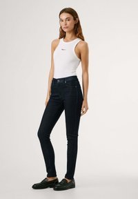 Bílý top bez rukávů s logem, v kombinaci s tmavě modrými skinny džínami vysoko nastavenými v pase. Džíny mají přední kapsy a hladkou texturu. Černé boty dokončují vzhled.