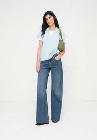 Ανοιχτό μπλε κοντομάνικο τοπ με V-λαιμόκοψη, συνδυασμένο με τζιν μπλε παντελόνι ψηλής μέσης και φαρδιάς γραμμής και μια μικρή πράσινη τσάντα χειρός.