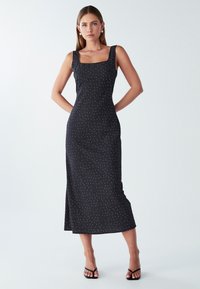 Žena stoji s rukama iza leđa, odjevena u crnu bezrukavnu midi haljinu s malim bijelim točkicama i crnim sandalama na petu.