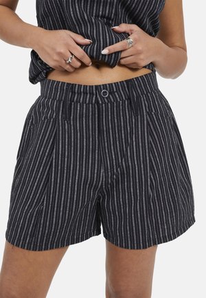 Femme portant un short noir à fines rayures et un haut assorti, soulevant le haut pour montrer le ventre, les mains tenant le tissu près de la taille.