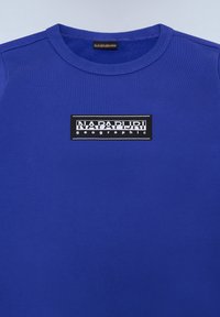 T-shirt azul de algodão com decote redondo e textura ribana; apresenta um patch de logo retangular preto com texto branco na parte da frente.