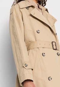 Beżowy trencz o podwójnym zapięciu, z dużymi okrągłymi guzikami, paskiem w talii i pagonami na ramionach. Gładka, trwała tekstura tkaniny.