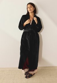 Schwarzer Samtrobe mit Schalkragen, Bindegürtel und seitlichen Schlitzen. Darunter ein bordeauxfarbenes Kleidungsstück mit Spitzenbesatz. Blumenschlappen runden den Look ab.