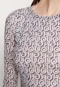 Długi rękaw przezroczysta bluzka z ogólnym wzorem splatających się liter w czerni na jasnoróżowym tle. Miękka, lekka tkanina.