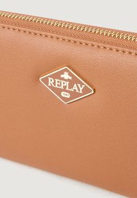 Portofel din piele maro deschis, cu o textură specială, având un fermoar auriu și un plăcuță cu logo în formă de diamant pe care scrie "REPLAY 1981."