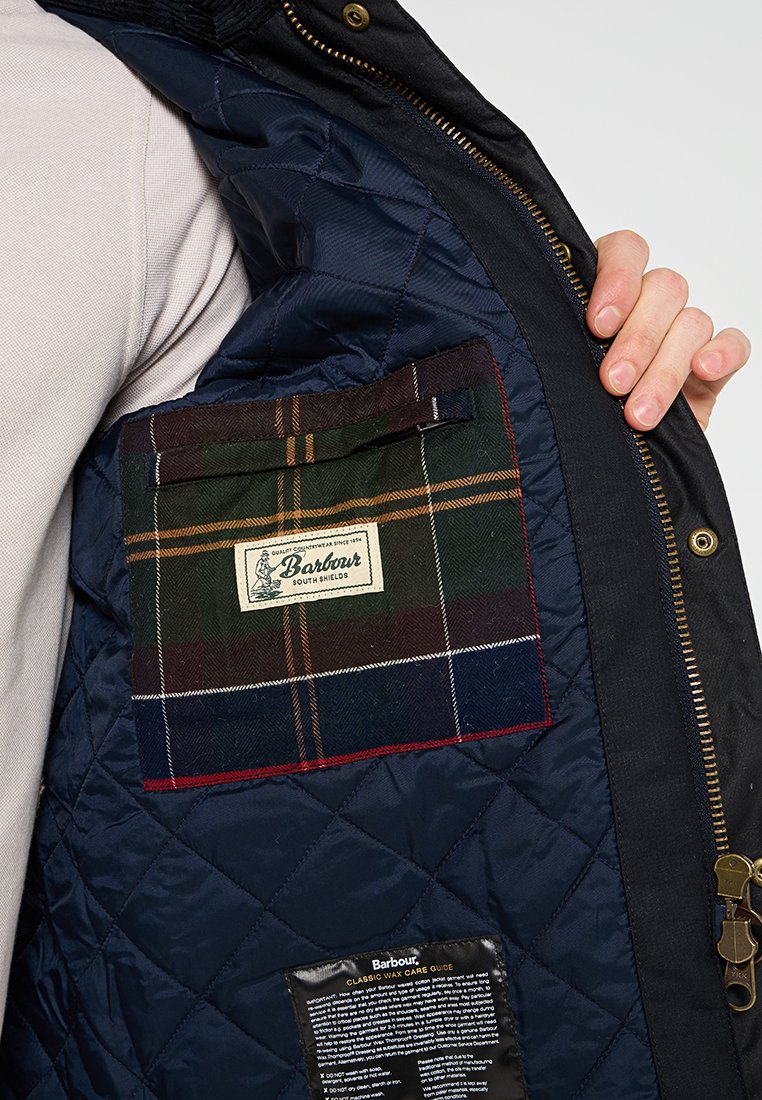 Main tenant une veste matelassée bleu marine ouverte montrant une poche intérieure à carreaux avec l'étiquette Barbour et l'étiquette d'entretien du tissu à l'intérieur.