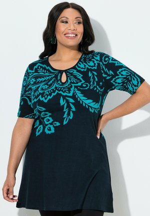 Lächelnde Frau mit dunklen Haaren trägt eine schwarze Tunika mit türkisfarbenen Blumenmustern und Schlitz am Ausschnitt, posiert mit einer Hand in der Hüfte.