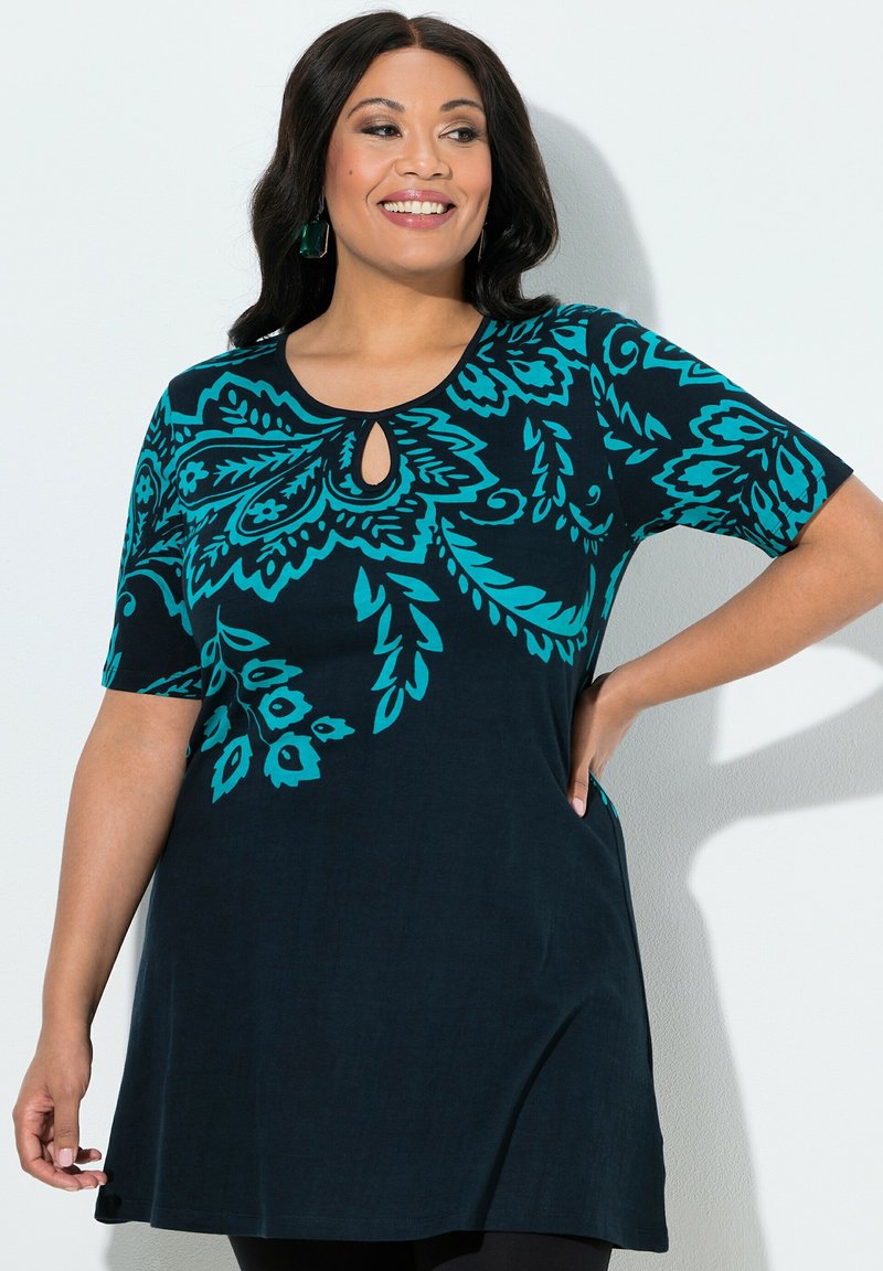 Smilende kvinde med mørkt hår iført en sort tunika med turkise blomsterprint og udskæring ved halsen, poserer med den ene hånd på hoften.
