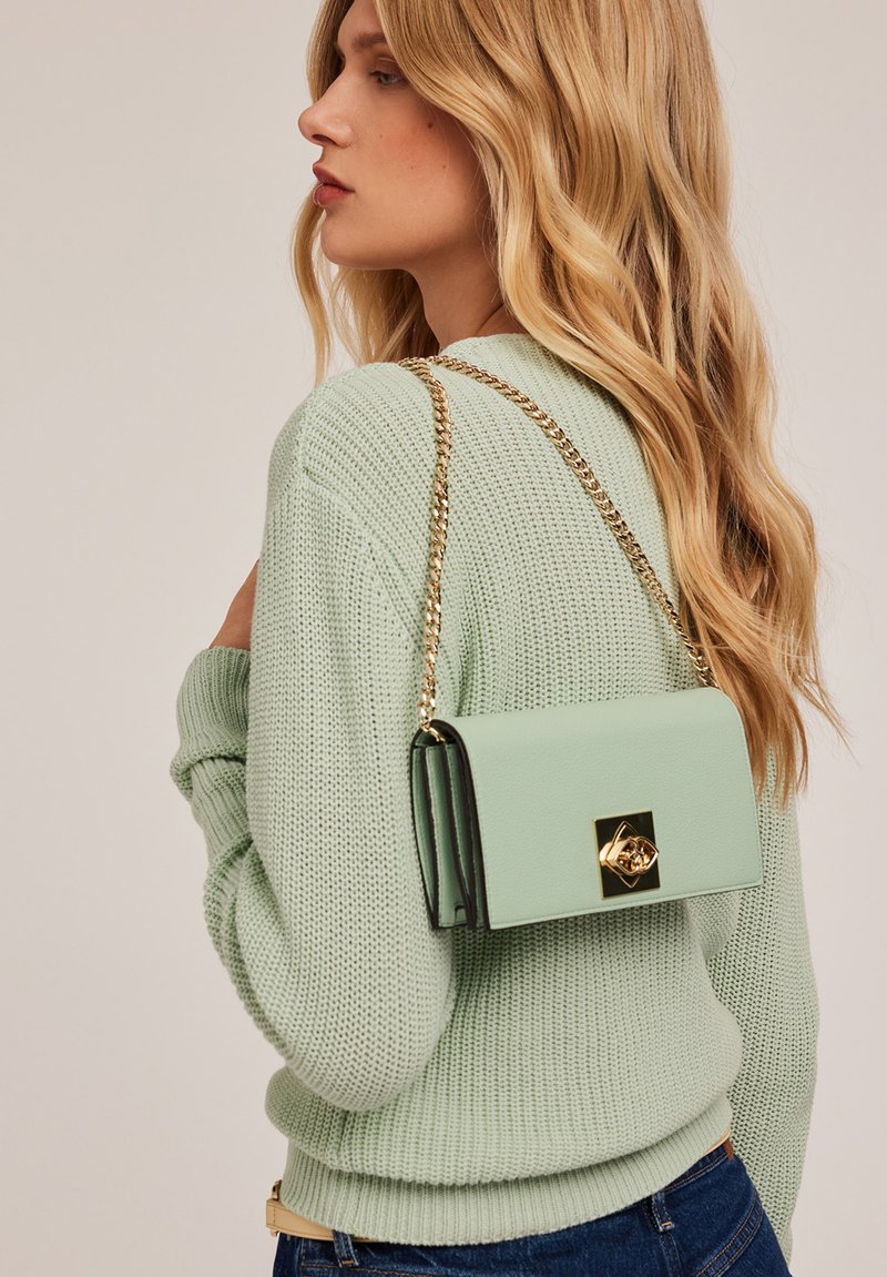 Bolso de piel verde menta con cadena dorada y ribetes negros, que presenta un diseño rectangular y un detalle de cierre decorativo.