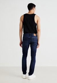 Jeans de mezclilla azul oscuro de corte ajustado, con bolsillos traseros y una etiqueta de cuero en la cinturilla. Llevados con una camiseta sin mangas negra y zapatillas blancas.