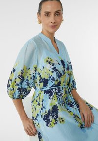 Himmelblaues Blumenkleid mit Puffärmeln, das gelbe und navyfarbene Muster aufweist. Bindung in der Taille und glattes, strukturiertes Material.