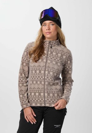Une femme porte une tenue de sports d’hiver : une polaire zippée à motifs, un pantalon de ski noir, un bonnet en maille et des lunettes de ski à verres réfléchissants.