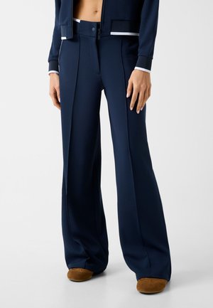 Frau trägt marineblaue, weit geschnittene, strukturierte Hosen und braune Wildlederschuhe, steht vor einem schlichten hellen Hintergrund.