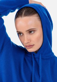 Hoodie bleu en maille avec capuche à cordon, poignets côtelés et texture douce, présenté avec une coupe décontractée et un design minimaliste.