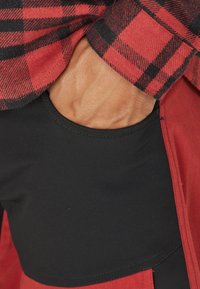 Röd och svart rutig skjorta med en kontrasterande svart ficka, som visar en slät textur och en figursydd design. Synliga sömdetaljer vid sömmarna.