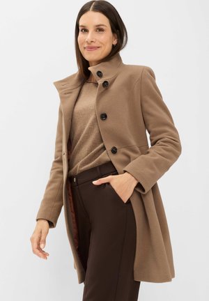 Frau trägt einen hellbraunen Wollmantel mit schwarzen Knöpfen, einen beigen Pullover und dunkelbraune Hose, steht mit einer Hand in der Tasche und lächelt.