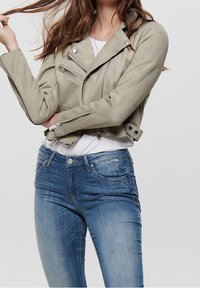 Grå ruskinds motorcykeljakke med sølvhardware, der har lynlåse og en tætsiddende pasform. Bæres over en hvid skjorte og parret med blå jeans.